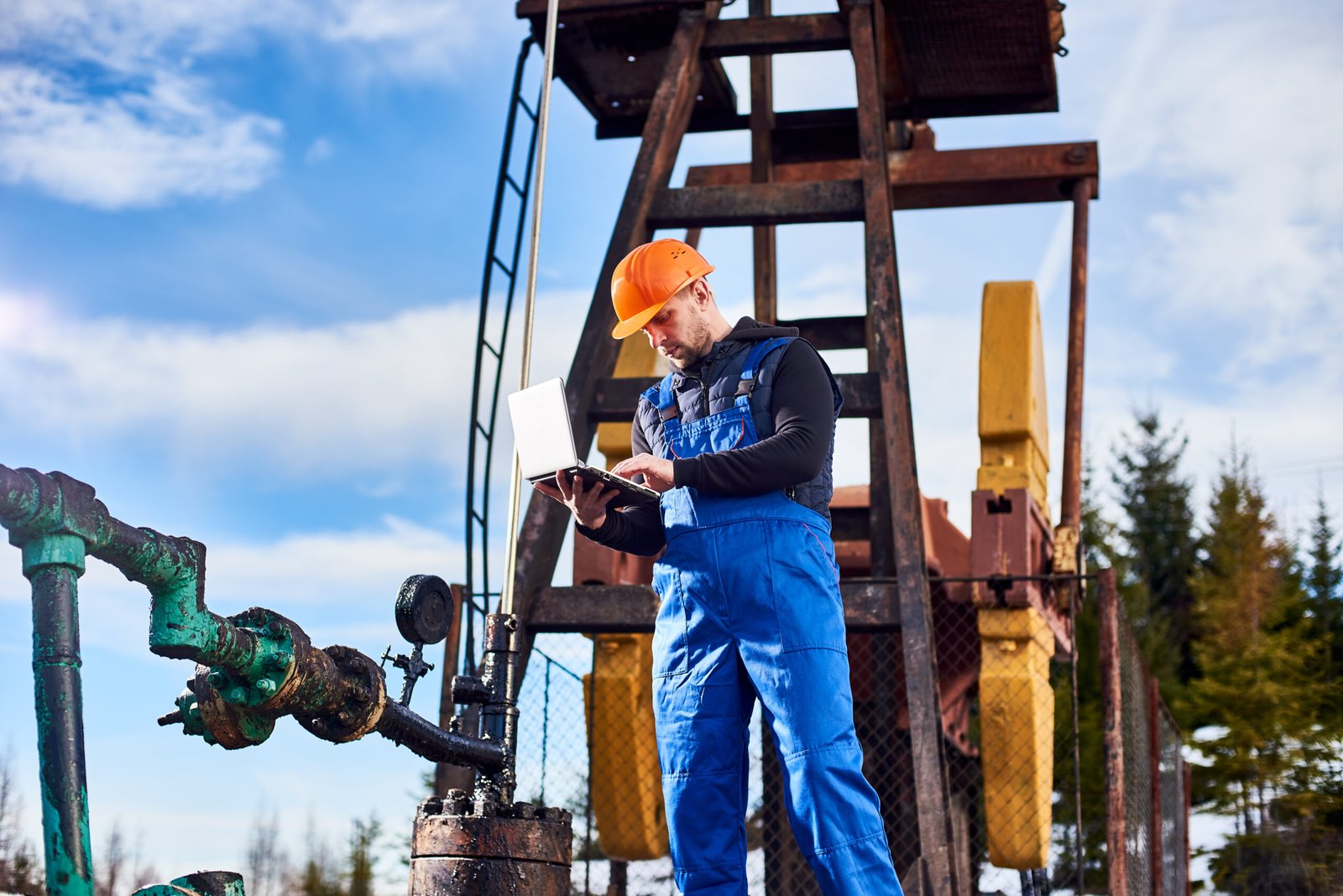 Engineer in work overalls and helmet checking oil pumping unit at oil field, with portable notebook. Male worker standing in front of oil well pump jack. Concept of petroleum industry, oil extraction.